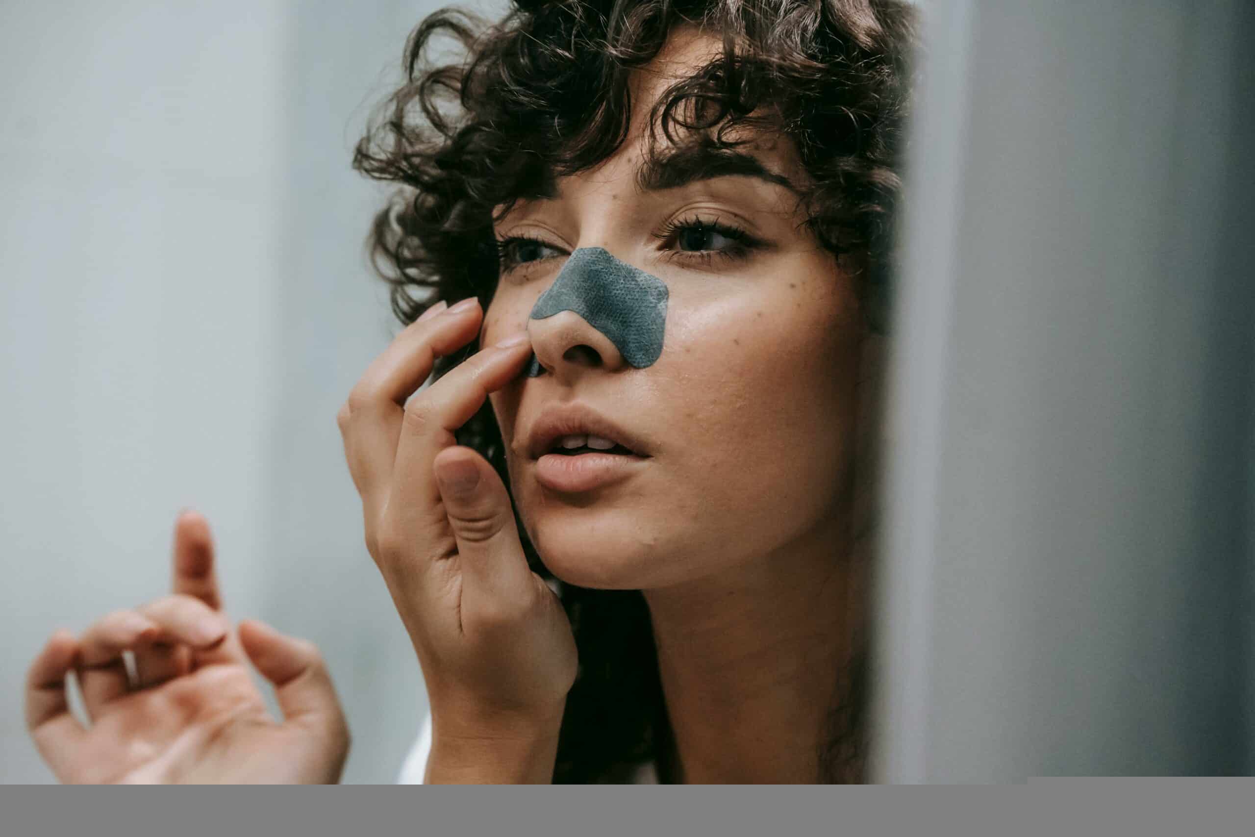 Mirror reflection of young female with curly hair putting nose strip mask on face in bathroom