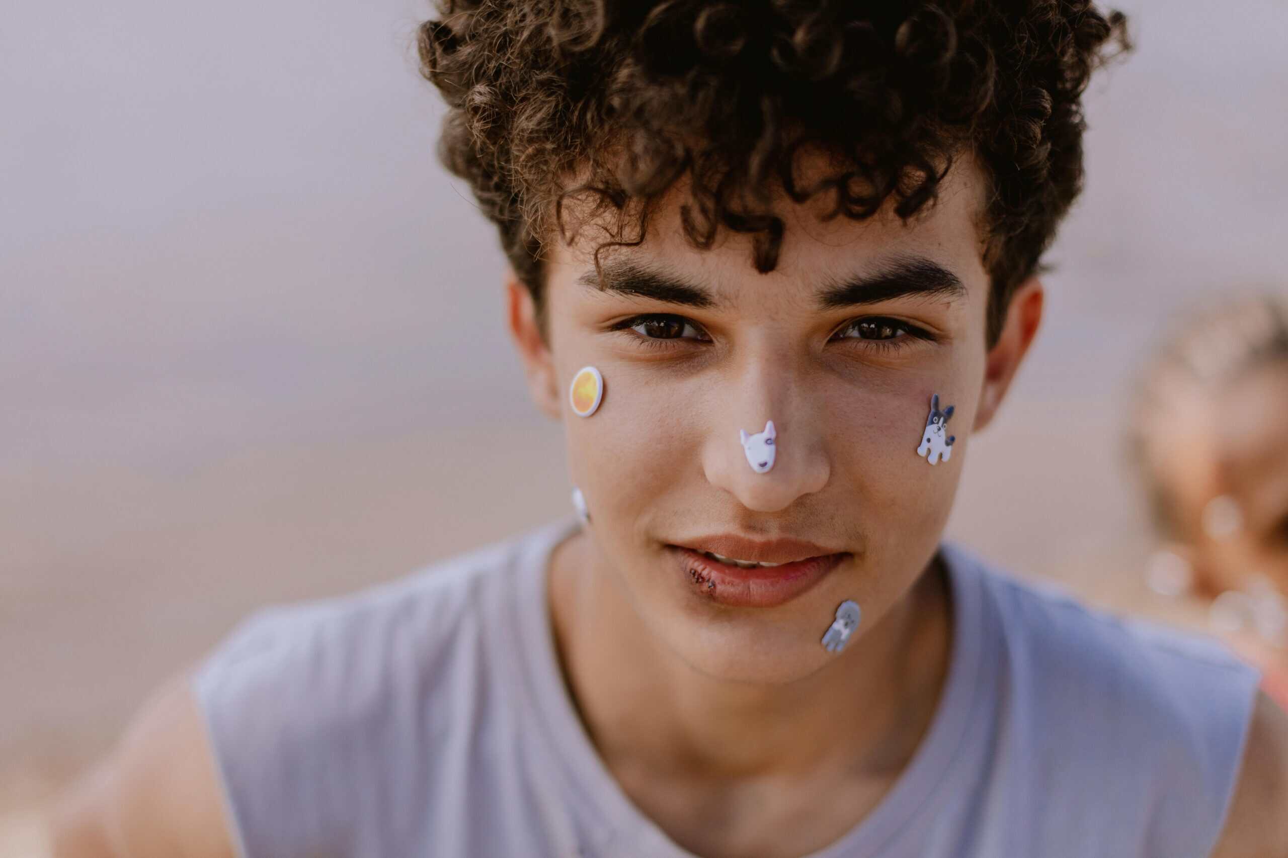 Close-up portrait of a teenage boy with playful stickers on his face, smiling outdoors.